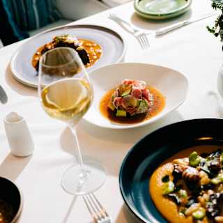 A table set with elegant crockery, including plated gourmet meals and a glass of white wine, on a white tablecloth in a bright, upmarket dining setting.