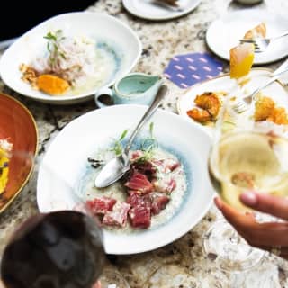 Guests raise glasses of red and white wine above a busy Tragaluz lunch, with a central plate of vibrant Nikkei tuna ceviche.