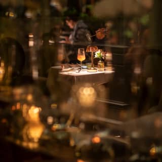 An orange cocktail sits in honeyed light on a dining table, seen through a haze of evening lights at Tragaluz restaurant.