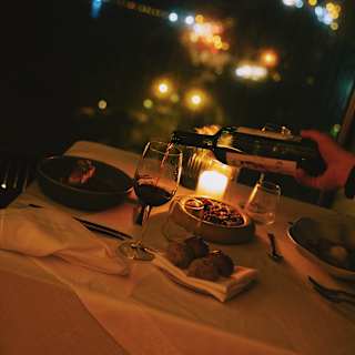A waiter tops up a glass of red wine at a candlelit table, viewed at an angle at night time, with a backdrop of city lights.