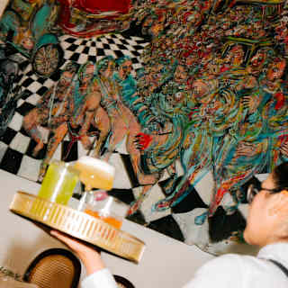 A waiter carrying a tray with drinks walks past a vividly coloured, abstract mural on a wall in a restaurant. The painting features dynamic, crowded scenes and a black-and-white chequered pattern.