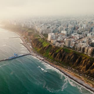 Aerial view of Lima buildings and green cliffs which overlook the beach circuit road and a jetty extending into a teal ocean.