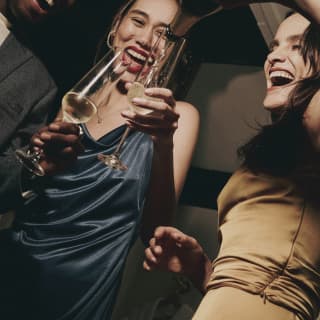 Low-angle shot of a woman in a gold dress laughing with a friend in a satin blue dress, as she chinks her champagne glass.