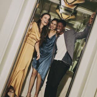 Angled shot of a woman in a gold dress standing with a friend in a blue dress and male companion in the open door of a lift.