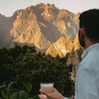 A man in a blue shirt stands with a drink on a viewing shady platform, looking out at the sun-capped contours of the Andes.