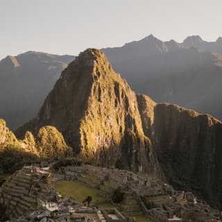 Green-gold with sun, Huayna Picchu looms over the shaded Machu Picchu ruins in a breathtaking aerial view of the citadel.