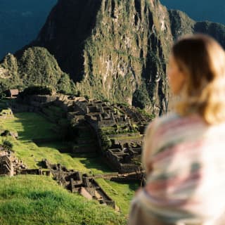 In soft-focus foreground and woman sits, gazing out over Machu Picchu, with the imposing, sun-kissed Huayna Picchu in sharp focus.