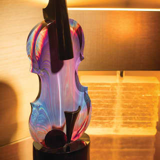 A small colourful glass sculpture of a cello on a podium stands on a polished sideboard, lit by a table lamp