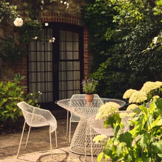 A terrace with white table and chairs by Emu is enclosed by lush foliage and Le Manoir's stone edifice, with climbing roses.