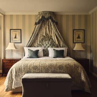 View of Hyacinth Suite's bed, regal with princess-style drapes, ornate padded headboard and furnishings in French greys.