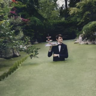 Surreal image of a waiter with a tray of afternoon tea cakes on his shoulder as he walks waist high through green pond weed.