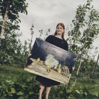 Standing in gardens, fallen apples at her feet, a woman with red hair, wearing black, holds a large still life of vegetables.