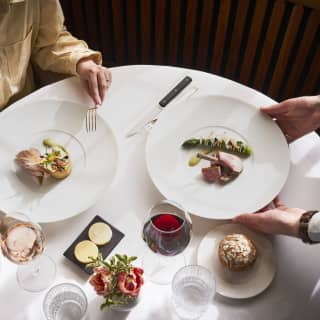 A waiter serves lamb cutlets with asparagus on a table for two, set with glass of rose, glass of red, bread rolls, butter and water