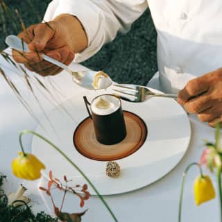 Close-up of an outdoor table as a chef holding a fork and spoon dips into the creamy froth of a café crème, seen from above.