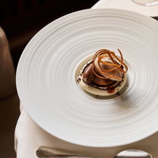 A coffee and cacao dessert with a chocolate quenelle and crisp garnish, served on a white dish with concentric grooves.