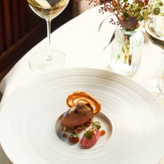 A plate of iced parfait of chicken liver with balsamic pear, herb petals and a crisp, at a table with white wine and flowers.