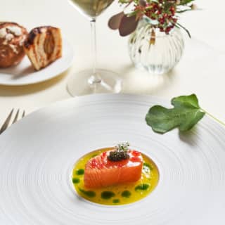 A white plate with a confit fillet of Loch Duart salmon, served in herb-split calamansi sauce, garnished with a fig leaf.