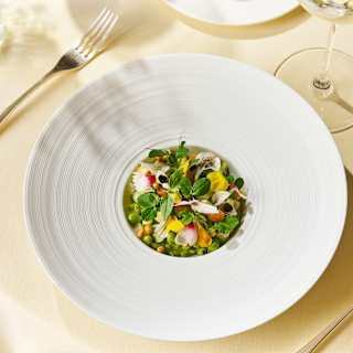 A wide-brimmed white bowl cradles a risotto of garden vegetables and herbs with vibrant bursts of green, yellow and pink.