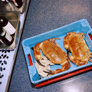 Two large, red-shelled brown crabs sit in the bottom of a blue crate on the kitchen floor next to a shelf of cooking wine.