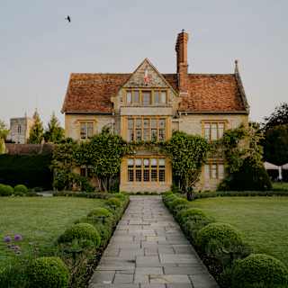 A bird soars above Le Manoir, seen at eye-level along a paved path flanked by lawn and shaped shrubs.