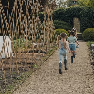 Three young girls in wellies run along a gravel path past bamboo cane pyramids awaiting the first tendrils of climbing beans