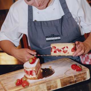 A female cookery-school student in a grey apron carefully lifts a slice of strawberry-infused cake from a chopping board.