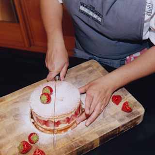 A guest slices a cake dusted with icing sugar and bordered with strawberry slices on a chopping board with five strawberries.