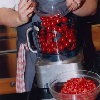 A man in a grey apron tips a glass bowl of cherry tomatoes into a food processor in a cookery class, seen from the neck down.