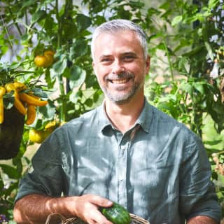 Acclaimed author and gardener, Rob Smith holds a basket of fresh cucumbers in a verdant kitchen garden dripping with produce.