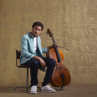 The seated young cellist, Sheku Kanneh-Mason, holds his cello and bow in his left hand as he looks off camera to the right