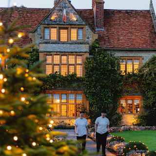 Two staff members walk on a path away from Le Manoir, whose windows glow with lights, seen past an illuminated garden fir.
