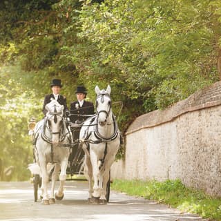 2 white horses pull an open carriage along the stone wall flanked road. 2 groomsmen wear top hats and white buttonholes