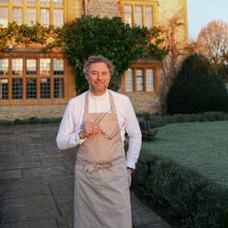 Chef Arnaud Donckele stands on a path in front of Le Manoir, smiling to camera and holding spectacles, face in late sun.
