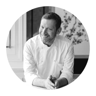 Black and white porthole portrait of Chef Patissier, Benoit Blin, smiling in chef whites with his hands together on a table.