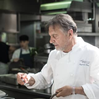 Side-view of Raymond Blanc in chef whites tasting a recipe from a silver spoon