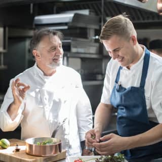 O chef Raymond Blanc sorrindo ao lado de outro chef na cozinha de um hotel