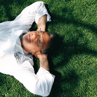 Chef Raymond Blanc, wearing glasses and chef whites, lies back on the grass with his arms behind his head, seen from above.