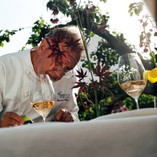 Low-angle image of Chef Raymond Blanc in spectacles as he reads at a table with two glasses of sparkling rosé in the garden.