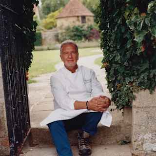 Raymond Blanc wears chef whites, blue trousers and boots as he poses for a photograph, sitting on a stone garden step.