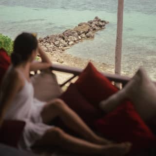 A woman relaxes on her terrace in soft-focus, gazing at the rocky coastal support that juts into the aquamarine sea below.