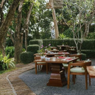 View along a row of five wooden dining tables on a gravelled area in a garden corner of Lai Thai circled by hedges and trees.