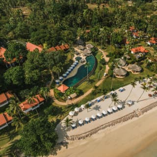 In an aerial view, loungers line a sandy terrace overlooking the sea with orange-roof residences and tropical pool behind.