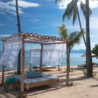 A swinging mattress of a simple wooden beach cabana provides shade on the sandy beach