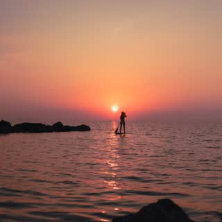 Silhouette of a woman paddle-boarding around dark rocks in a sea glowing with the reflection of the red setting sun.