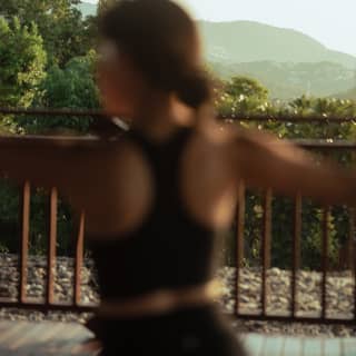 A woman in a black top does yoga in soft-focus foreground at the pavilion, with green mountain views, hazy in sunshine.