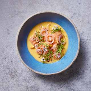 A blue bowl filled with creamy yellow soup, garnished with thinly sliced vegetables, fresh herbs, and edible flowers, sits on a light grey textured surface.