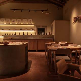 A warmly lit, minimalist restaurant interior with wooden tables and chairs, a central round table, shelves with art and bottles, and a textured wall decoration resembling a branch.