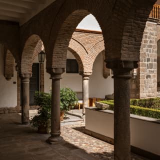 View along a shadowy arcade with glimpses of a sunny courtyard garden and perpendicular arcade made with stone and brick.