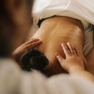 A person receives a back massage while lying face down on a massage table, partially covered by a white sheet. The masseuse's hands are pressing gently on their upper back.