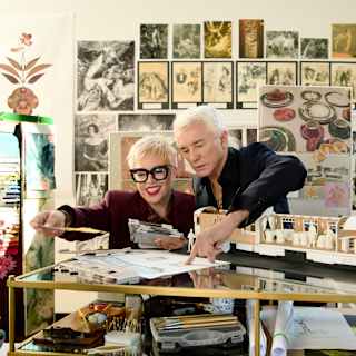 Two people stand smiling in front of a colourful wall mural featuring stylised flowers and dancing figures in whimsical costumes. One person has short white hair, the other has short blonde hair and wears sunglasses.